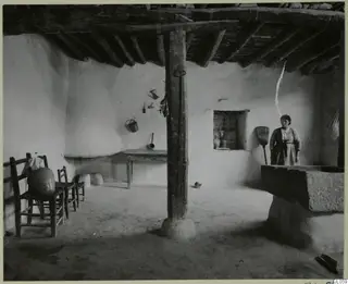 Interior of a whitewashed village house with a timber-beamed ceiling, a woman standing by a broom at right.