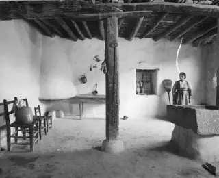 Interior of a whitewashed village room with a timber-beamed ceiling and a woman standing by a broom at the right.
