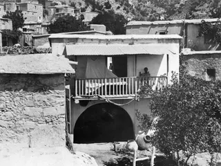 Buildings, trees and a man at Mandres, with a whitewashed house, arched passage and balcony in a hillside village.
