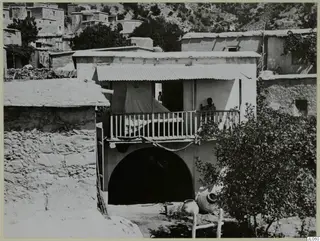 Two-storey building with an arched ground floor and a wooden balcony at Mandres, with a man standing on the balcony.
