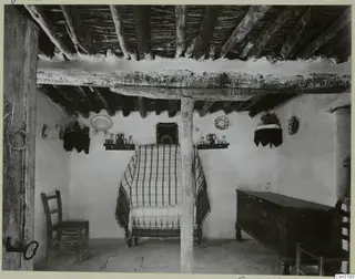 Interior of a rural room with a timber-beamed ceiling, a table covered by a striped cloth, and a painted chest against the wall.