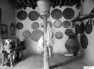Interior of a house with round flat trays hung on the plastered walls and a large jar standing on the floor.