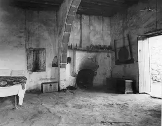 Interior of a plastered room with a bed, a fireplace shelf holding bottles, a wooden chest and an open door