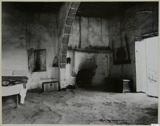 Interior of a plastered room with a shelf of bottles above a hearth, a bed at left and a wooden chest on the floor