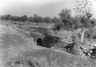 Stone arch of a vault or culvert crossing a dry channel beside a dirt track, with trees and flat fields behind