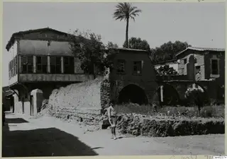 Street in Nicosia with a two-storey timber-fronted house, arched stone walls, a tree and a standing man in light clothing.