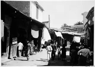Street in Nicosia lined with shopfronts, white cloths hung over the awnings, and several people walking along the roadway