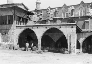 Sacks and two standing men in front of an arcaded stone building in Nicosia