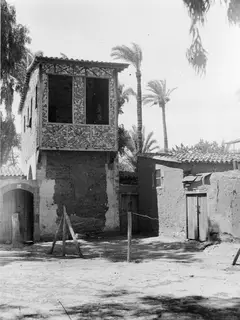 Two-storey house with a decorated timber upper balcony and a mud-brick building beside it, with palm trees behind, in Nicosia.