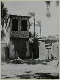 Two-storey building with a timber upper floor of patterned panels, palm trees and a mud-brick annexe beside it, in Nicosia