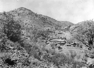 Village of Lazania with flat-roofed houses clustered on a terraced hillside, seen from a rocky slope with trees