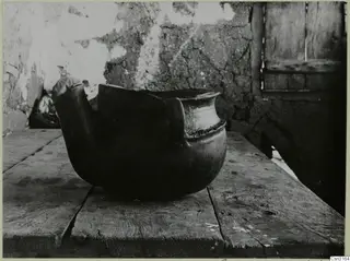 Large round-bodied clay vessel with a handle and broken rim, standing on a wooden table in Klirou