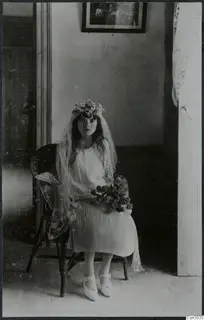 Young woman seated in a wicker armchair, wearing a flower crown and veil, holding a small bouquet, at Lapithos.