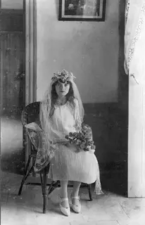 Young woman with a veil and flower wreath seated in a wicker chair, holding a bouquet, at Lapithos