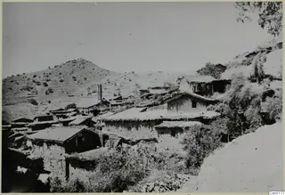 Village of Lazania on a hillside, with stone houses with tiled and flat roofs, a slender tower and trees.