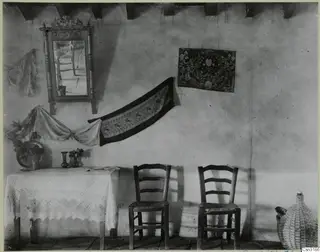Interior with a carved mirror, embroidered textiles, a lace-covered table and two rush-seated chairs against a whitewashed wall