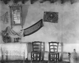 Interior with a lace-covered table, two rush-seated chairs, a framed mirror and embroidered textiles on a whitewashed wall