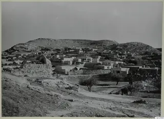 Galinoporni village of whitewashed houses on a slope below bare hills, with a stone wall and trees in the foreground.