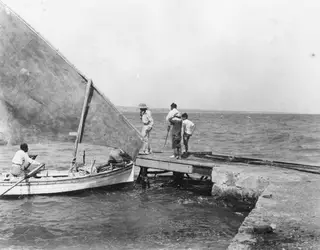Sailing boat with a large sail alongside a wooden jetty, with several people in the boat and on the jetty