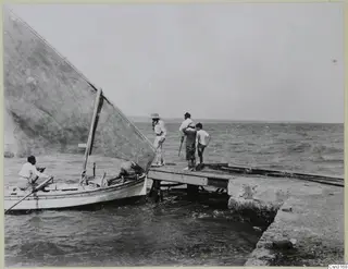 Small sailing boat with a large sail alongside a stone jetty, with several people standing and sitting nearby by the sea.