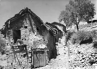 Stone houses along a narrow stony village street in Lazania, with a wooden ladder leaning against a house wall.