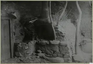 Stone oven with a dome-shaped opening in a stone-walled room, with a wooden door at left and wooden poles against the wall.