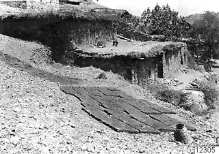 Stone-walled building at Lazania with a flat roof, and flat dark pieces laid out on the ground in front of it.