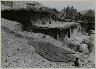 Stone house with a grass-covered flat roof and rows of flat cakes laid out on a stony slope at Lazania