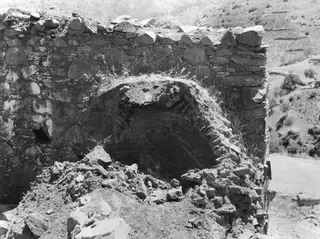 Stone oven with an arched opening set into a rubble-stone wall, with fallen stones and soil in front.