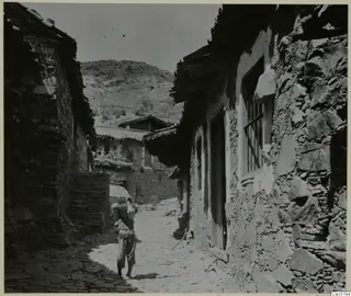 Stone-built village street in Lazania, with a figure carrying a load on the head and a hillside behind the houses.