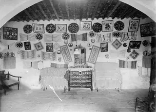Interior of a cottage at Milia with embroidered wall hangings, framed pictures, two beds and a carved chest of drawers