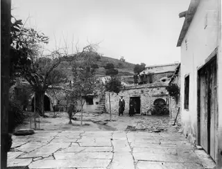 Stone-walled courtyard with flagstone paving, a domed oven recess in the wall, and people standing by a doorway