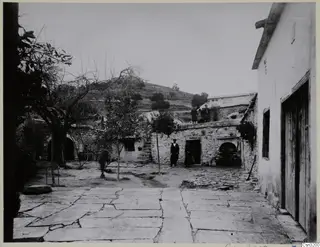 Stone-paved courtyard with a rubble-stone wall, a possible oven recess, and people standing on the wall top