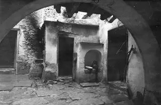 Stone-paved courtyard under an arch, with a whitewashed wall holding an arched niche that contains a bowl and a jug