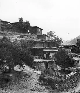 Stone and mud-roofed village houses built up a hillside among trees, with mountains behind.