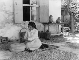 Woman kneeling to turn a hand mill outside a whitewashed house, with jugs on a table behind her.