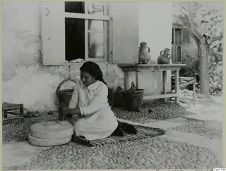 Woman grinding grain with a hand mill on a cobbled yard, with baskets and jugs beside a house wall