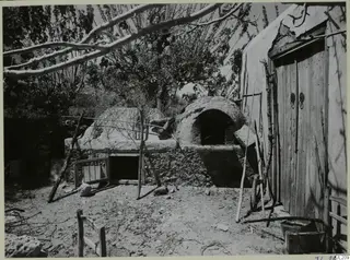 Domed clay oven with an arched opening in a dirt yard, beside a plastered building with wooden doors
