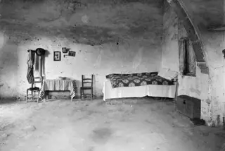 Interior of a whitewashed room with a bed, a table with a striped cloth, two chairs and a wooden chest.