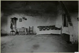 Interior of a whitewashed room with a bed, two wooden chairs and a table covered with striped cloth