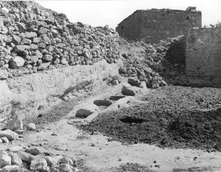 Stone feeding troughs cut into a pale ledge beside a rough stone wall at Galinoporni, with a ruined stone building behind.