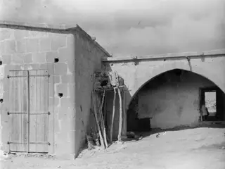 Stone building with a wooden plank door and an arched opening, with wooden poles leaning against the wall.