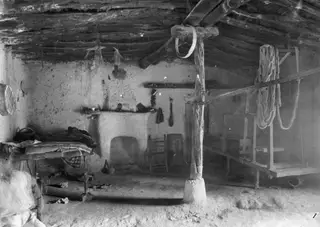 Interior of a plastered room with a branch roof, a wooden loom on the right and a wooden bed on the left.
