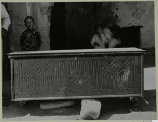 Carved clothes chest, so-called sendudzin, standing outdoors in front of a plastered wall with children nearby.
