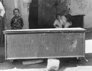 Carved wooden clothes chest standing on stone blocks, with children behind it against a plastered wall