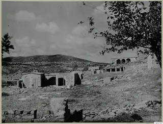 Stone buildings with arched openings and low dry-stone walls on a stony slope, with a bare hill behind, in Cyprus