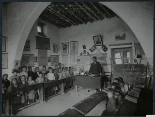Teacher and pupils in a whitewashed classroom at Lapithos, with wall charts and a chalkboard