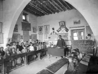 Teacher standing at a desk in an arched whitewashed classroom, with rows of children on benches, Lapithos