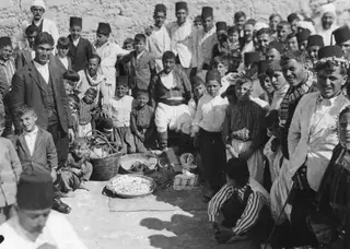 Crowd of men, boys and a woman gathered around baskets and a tray of small pale items at Galinoporni.