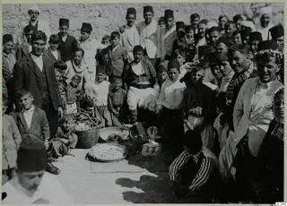 Crowd of men, boys and a woman in fezzes and jackets gathered around baskets and a food tray at Galinoporni.